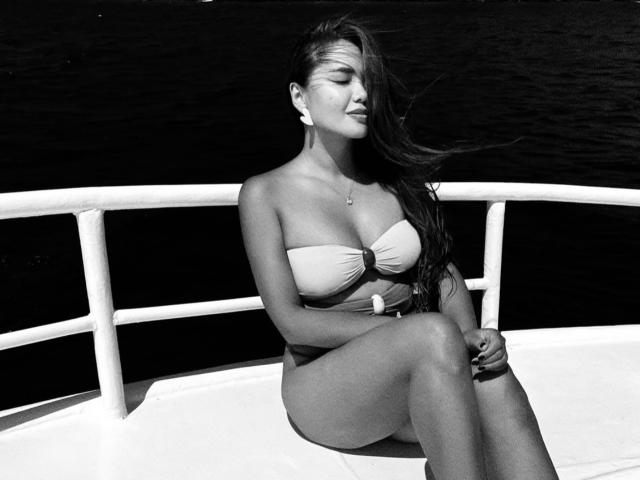Foto em preto e branco de KetrinMagicx com cabelos longos e escuros, olhos fechados, usando um biquíni sem alças, sentada em um barco, pernas cruzadas, braços relaxados, mostrando seus seios e nádegas..