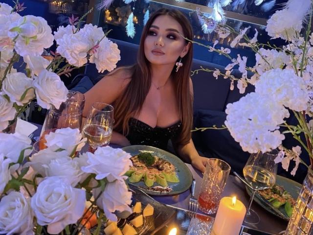 KetrinMagicx, con cabello largo y castaño, busto grande, vestida con un vestido negro brillante, sentada a una mesa con rosas blancas, velas y cristalería, mirando hacia arriba con una sonrisa sutil..