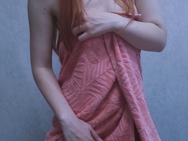 SheritaChieng with pale skin and red hair, holding a pink towel over her bare tits and pussy, covering her chest and lower body with her right hand.