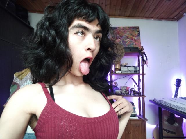 FridaBiscuit with curly black hair, brown eyes, and open mouth sticking out her tongue, wearing a red ribbed tank top, showing her small tits.