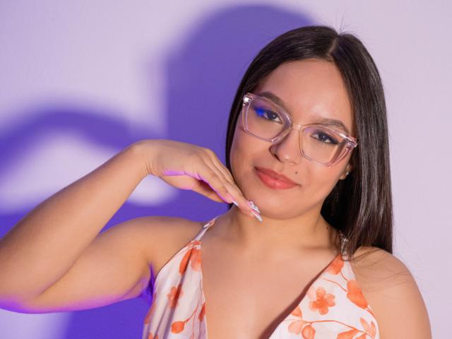 CatalinaPorto with glasses, long dark hair, smiling, holding her chin, wearing a floral dress, showing her tits, light skin, pink nails, purple shadow on her face.