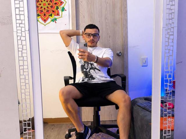 LianFerrer, sitting on a chair, legs spread, taking a mirror selfie, wearing glasses, white tee, black shorts, sneakers, wristbands, wooden door, colorful art behind, blue light.
