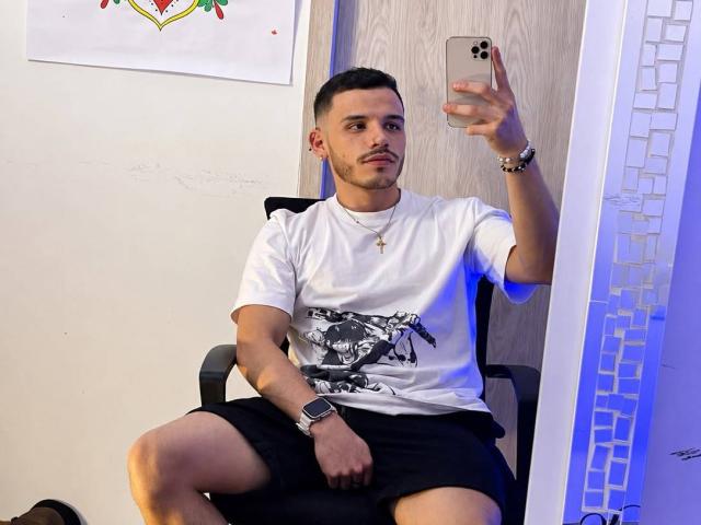 LianFerrer, with short dark hair, white t-shirt, black shorts, and a watch, takes a selfie in a mirror. He's sitting, holding a gold phone, with a serious expression.