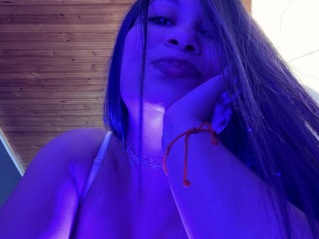 KendallBrzz with blue-lit skin, long brown hair, white bra, lace choker, hand on cheek, orange bracelet, subtle smile, soft makeup, low-angle shot, looking down at camera.