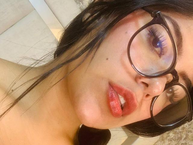 CatalinaReiyna with red lipstick, glossy lips, black glasses, blue eyes, and dark hair, showing a small nose piercing, looking at the camera with a slight open-mouth expression.