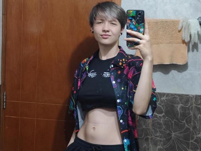LissyLuxe with short brown hair, black crop top showing small tits, colorful floral robe, black pants, holding phone in mirror, smiling, towel on wall, wooden door.