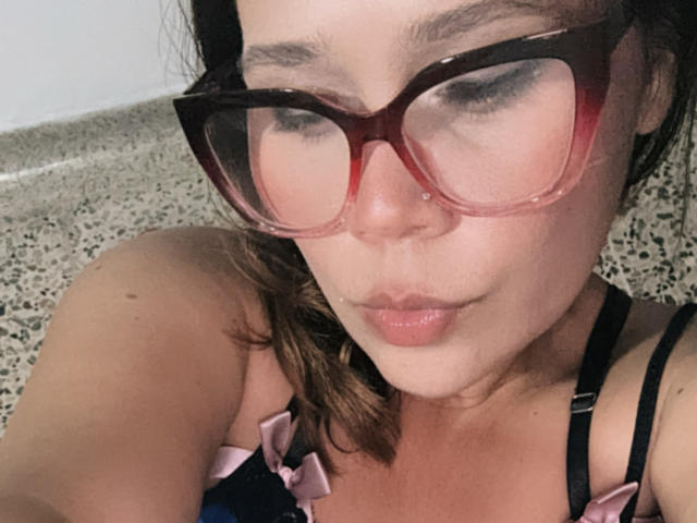 SophiaQueens with red glasses, pouty lips, and brown wavy hair, wearing a pink and black strap dress, looking down, slight nose piercing, freckles, and light skin.