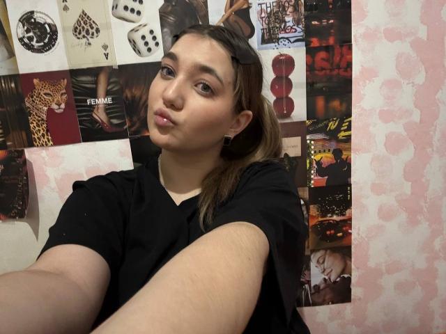MichikoVivona, met volgezwollen lippen, bruin haar in een paardenstaart, draagt een zwarte top, heeft subtiele make-up, een zwarte zonnebril op haar hoofd, een roze behang achter zich, in een selfie-stijl, een casual uitstraling, met een gemiddelde borstgrootte..