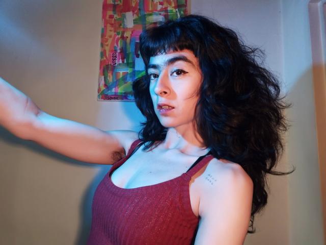 FridaBiscuit with wavy black hair, dark eyebrows, and a nose ring, wearing a red tank top, exposing her armpit hair, and small tattoo on her right shoulder.