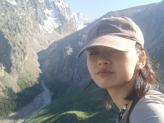 EmiliaBrackemyre with brown cap, brown hair, and light skin, showing a serious face, wearing a gray shirt, in a mountain landscape with rocky cliffs and green valleys.