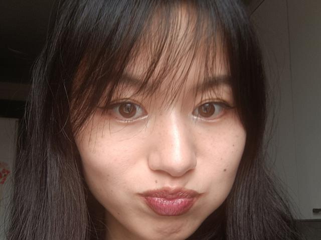 EmiliaBrackemyre's face with black straight hair, brown eyes, shiny pink lips, and a small mole on her left cheek, looking straight at the camera.