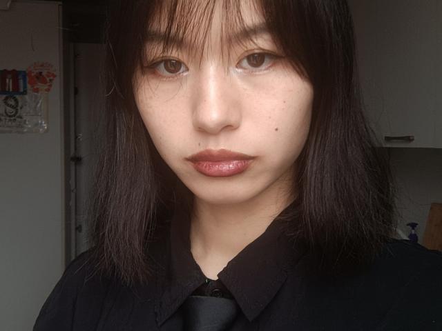 EmiliaBrackemyre with shiny pink lips, black shirt, black necktie, medium-length brown hair, bangs, light skin, brown eyes, small moles on face, slightly parted glossy lips, serious expression.
