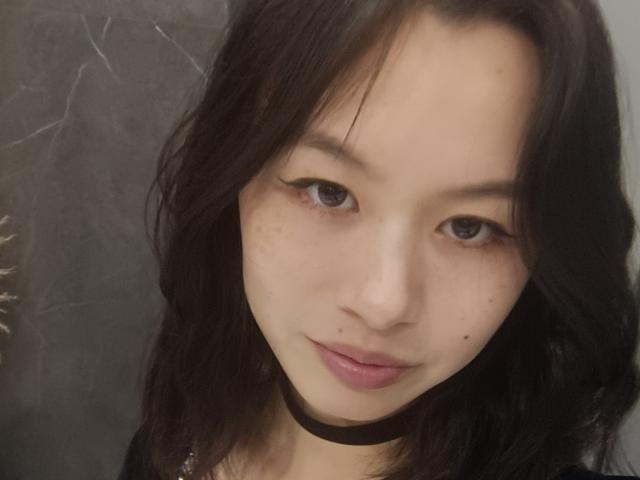 EmiliaBrackemyre has dark wavy hair, light skin, freckles, black choker, and a small beauty mark on her right cheek. She's looking at the camera with a slight smile.