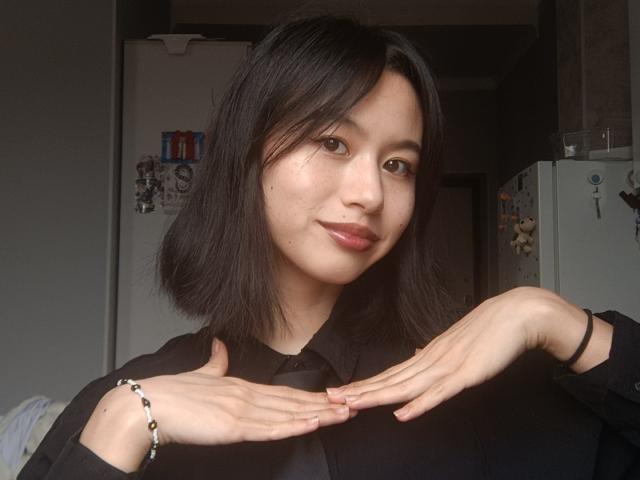 EmiliaBrackemyre with black shoulder-length hair, smiling, wearing a black shirt, hands gently touching chest, pink lipstick, bracelet on right wrist, subtle moles on face, soft natural light.