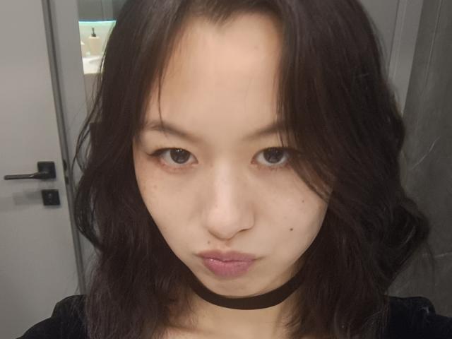 EmiliaBrackemyre with dark, wavy hair, pale skin, small nose, brown eyes, black choker, and pouty lips, staring into camera, showing a few small moles on her face.