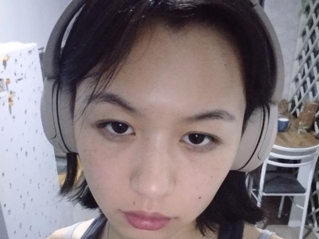 EmiliaBrackemyre has a small nose, dark eyes, and black hair, wearing grey headphones, with a neutral expression, and a small mole on her right cheek.