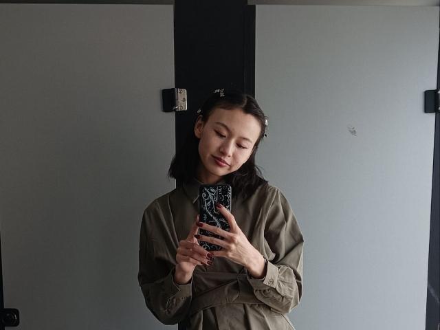 EmiliaBrackemyre with short black hair, green shirt, holding phone with black patterned case, smiling, eyes closed, standing in front of gray and black wall.