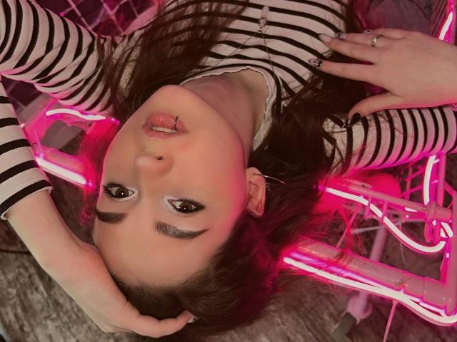 AndraAnestos with long brown hair, striped shirt, and pink neon lights, has a lip ring and hand on head, looking up with half-lidded eyes and pink-tinted face.