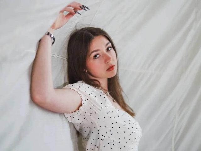 AndraAnestos with long brown hair, white polka-dot shirt, and red lips, lying on white fabric, arm raised, looking at camera, wearing a black bracelet, with long nails.