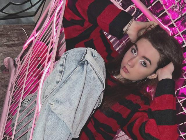AndraAnestos with dark brown hair, wearing a red and black striped sweater, and faded blue jeans, lying on a pink wire chair, hands in her hair, looking at the camera.