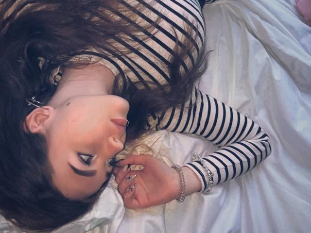 AndraAnestos is lying on a bed, tongue out, with striped black and white shirt, long brown hair, and nose ring, looking up, hand near mouth, white bedsheet.