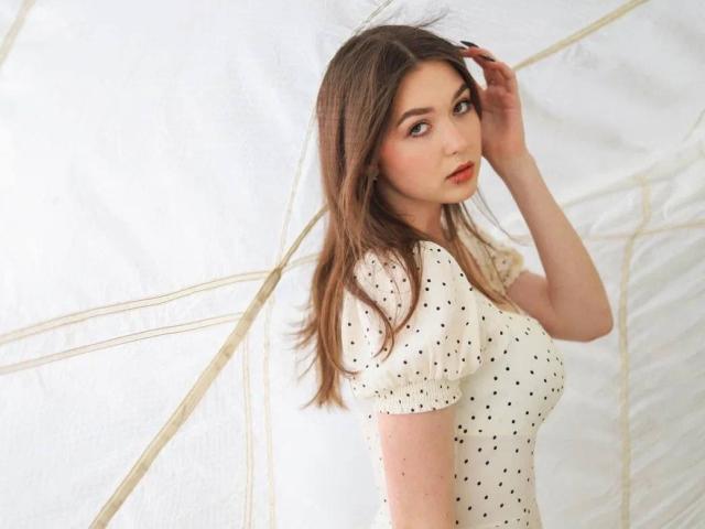 AndraAnestos with brown hair, wearing a white polka dot top, looking over shoulder, hand in hair, light skin, soft makeup, subtle red lipstick, gentle expression, standing slightly turned.