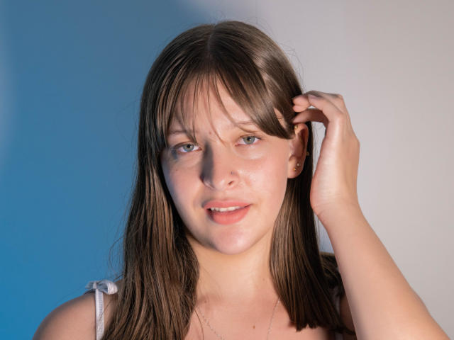 LiaMilan has straight brown hair, blue eyes, and fair skin with a subtle smile. She touches her hair with her right hand, wearing a white strap top.
