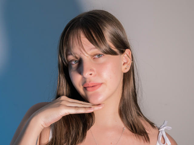 LiaMilan with brown hair, blue eyes, and pink lips, touching her chin, wearing a white strap top, looking at the camera with a slight smile, soft sunlight on her face.