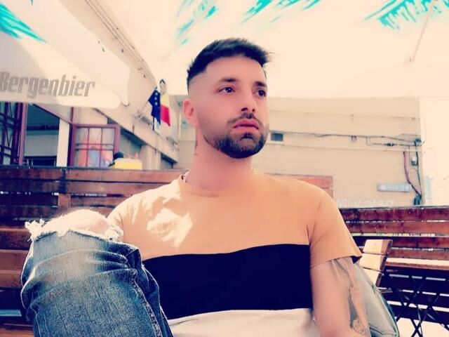 TommyDum with a trimmed beard and short haircut, wearing a black and white striped shirt, sitting with ripped jeans, tattoo on right arm, looking serious, outdoor setting.