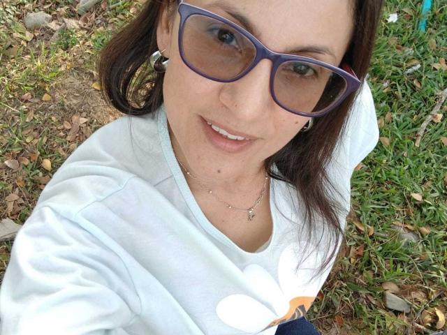 SolaraDubois with purple sunglasses, white shirt, and necklace, smiling, showing a small beauty mark on her chin, in a grassy outdoor setting.