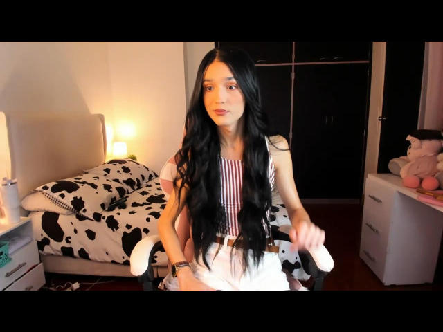 SophieCornets with long black hair, sitting on a chair, wearing a striped dress, holding her white panties, in a bedroom with a cow-pattern bed and pink stuffed toy.