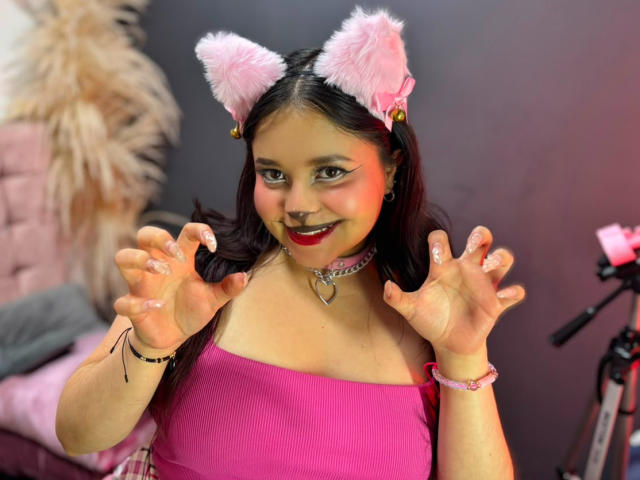 SamyMancini with pink furry cat ears, pink top, and glitter on hands, smiling, showing claws, pink choker, pink bracelet, and nose ring, in a cute, playful pose.