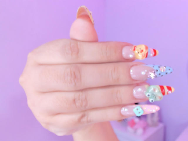 MarlaBriard's hand with long, colorful, and shiny nails featuring different designs like candy stripes, polka dots, and tiny eyes on a purple background.