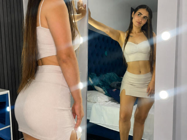 Orianafenix, avec de longs cheveux bruns, portant un haut court rose côtelé et une jupe, montrant ses fesses dans le miroir, debout dans une pièce avec un lit derrière elle..