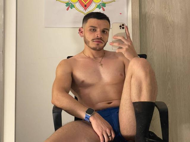 LianFerrer, shirtless, muscular, blue shorts, black socks, holding iPhone, cross necklace, wrist watch, short dark hair, light skin, seated, leg up, mirror selfie, wooden door, decorative wall art.