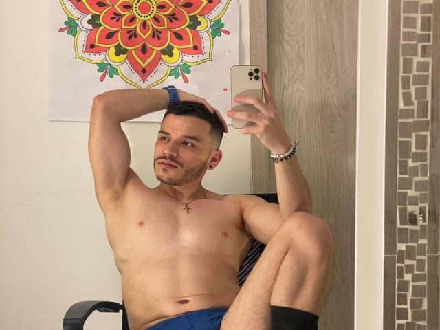 LianFerrer, shirtless, muscular, with a beard, taking a mirror selfie, blue shorts, black socks, cross necklace, bracelet, wood door, colorful mandala poster, holding iPhone, right arm raised.