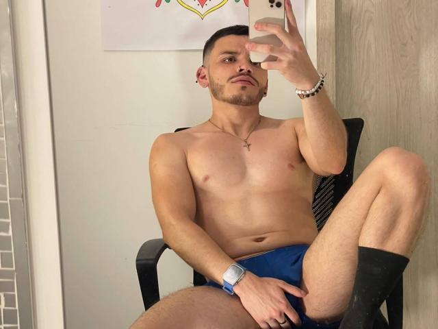 LianFerrer, shirtless, wearing blue underwear, black socks, taking a mirror selfie with his right hand, cross necklace, bracelet, and a watch, sitting with one leg up.