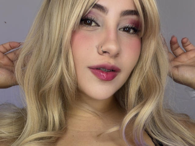 LorensDouce tem cabelo loiro, batom rosa e delineador escuro com sombra rosa. Ela está usando um piercing no nariz e tem as mãos perto da cabeça, olhando para a câmera..