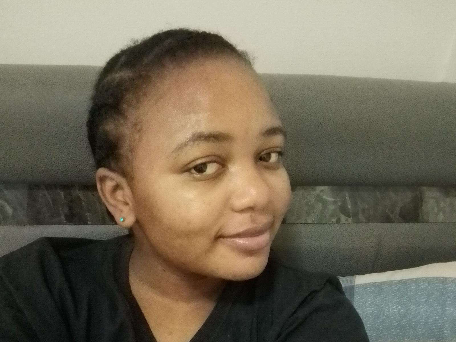 Sheilax, avec un petit sourire, porte une chemise noire, a les cheveux courts tressés, de petites boucles d'oreilles et un visage légèrement gonflé, et regarde sur le côté. C'est une femme à la peau foncée..