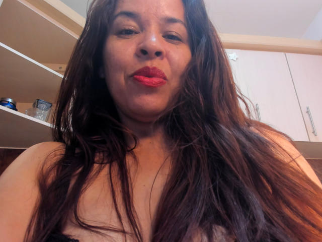 LizethBells has long brown hair, red lipstick, and a slight smile. She's topless with visible boobs and a light skin tone, in a kitchen with white cabinets.