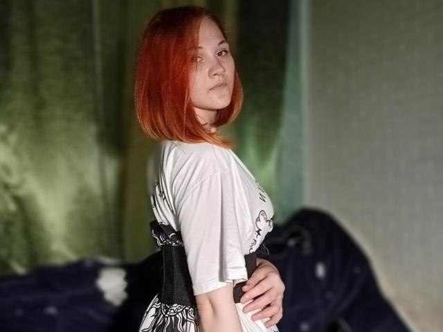 CarissaCariddi with red bob, white tee, black patterned dress, holding belly, green curtains, standing sideways, looking over shoulder, slightly parted lips, soft light, casual pose.