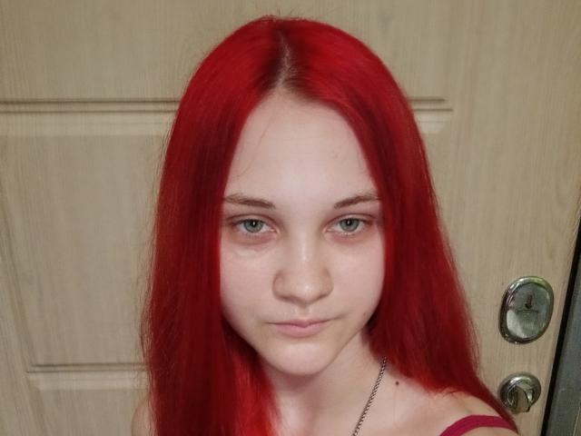 CarissaCariddi with red hair, pale skin, green eyes, and a subtle smile, showing her face and shoulders, wearing a red strap. Her pussy is not visible.
