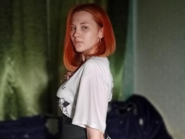 CarissaCariddi with red hair, white tee, and gray dress, standing side-on, looking at camera, small earrings, green curtain background, subtle smile, soft lighting.