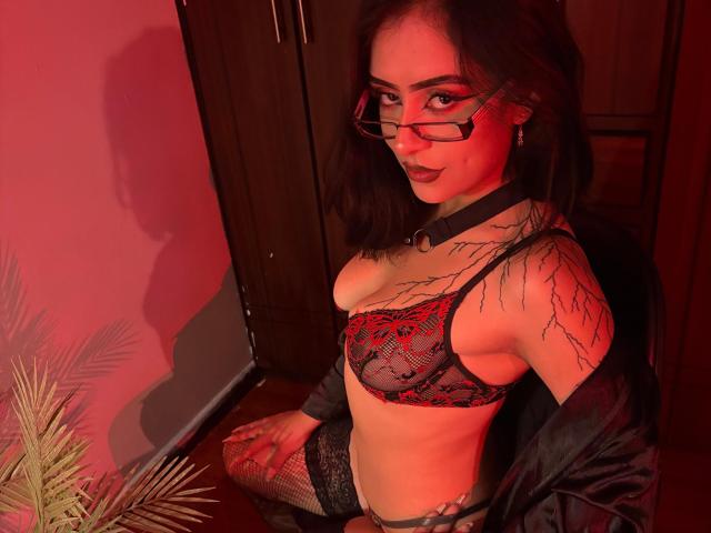 QueenMissty with red-laced black bra, tattoos on shoulders, black choker, glasses, dark lipstick, and black lace stockings, sitting in red light, black jacket off, looking at camera.