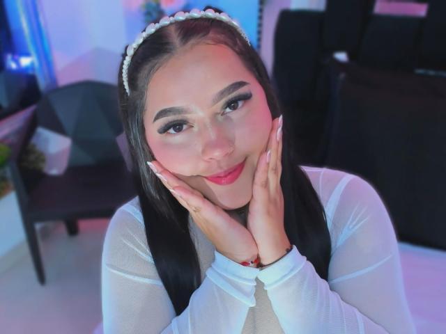CandasLeroy with dark hair, white headband, and sheer white top, hands on cheeks, smiling, pink lipstick, and long eyelashes, in a softly lit room.