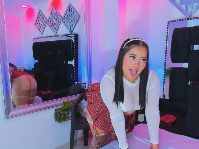 CandasLeroy, with black hair and white headband, wearing a sheer top, plaid skirt, and fishnet stockings, is on all fours, tongue out, with a reflection of her ass.