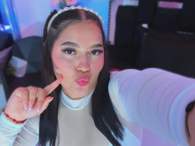 CandasLeroy, with pouty pink lips and finger on cheek, has long black hair, white headband, and white top, making a cute face in a selfie.