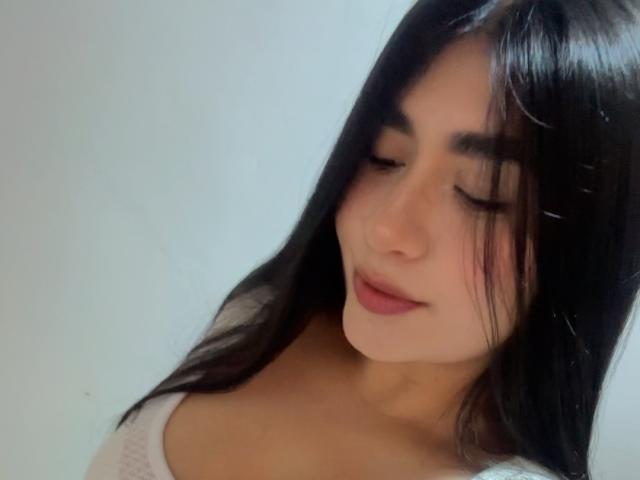 MiaDuboisX has long black hair, closed eyes, and a slight smile. She's wearing a white top with a low neckline, showing her cleavage. Her skin is light.