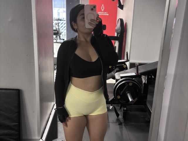 MiaDuboisX with tan skin, black sports bra, yellow shorts, black zip-up jacket, and black fingerless gloves, taking a mirror selfie in a gym. Her tits are visible.