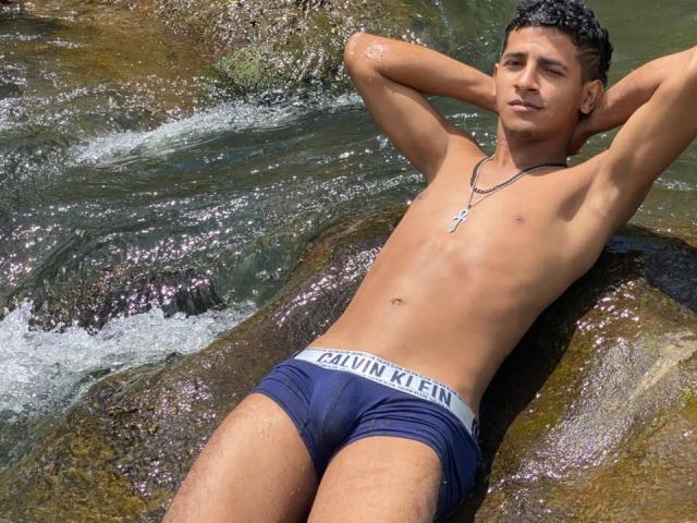 DannyBigCockx, shirtless, with hands behind head, wearing navy Calvin Klein briefs, lying on rocky shore, water flowing, necklace with cross, short dark hair, relaxed, confident expression.